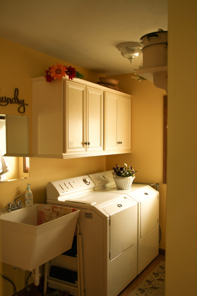 Foto på en mellanstor vintage linjär tvättstuga enbart för tvätt, med en allbänk, luckor med upphöjd panel, vita skåp, gula väggar, laminatgolv, en tvättmaskin och torktumlare bredvid varandra och brunt golv