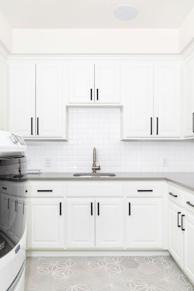 Bild på en mellanstor vintage grå u-formad grått tvättstuga enbart för tvätt, med en allbänk, skåp i shakerstil, vita skåp, bänkskiva i kvarts, vitt stänkskydd, stänkskydd i porslinskakel, vita väggar, betonggolv, en tvättmaskin och torktumlare bredvid varandra och grått golv