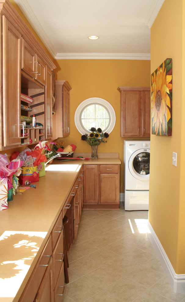 他の地域にあるトラディショナルスタイルのおしゃれな家事室 (L型、ドロップインシンク、落し込みパネル扉のキャビネット、中間色木目調キャビネット、左右配置の洗濯機・乾燥機、ベージュの床、ベージュのキッチンカウンター、オレンジの壁) の写真