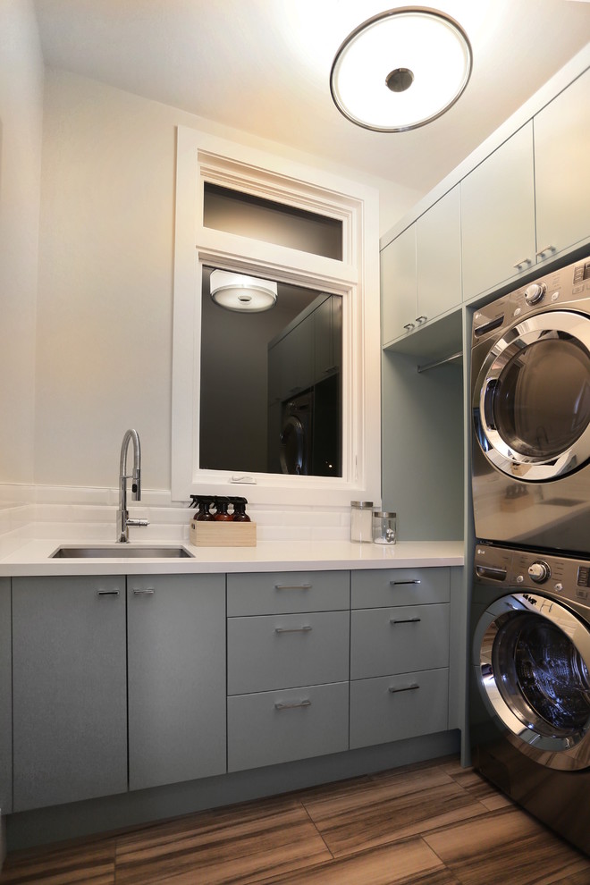 Inspiration for a mid-sized contemporary single-wall light wood floor dedicated laundry room remodel in Vancouver with a drop-in sink, flat-panel cabinets, white walls, a stacked washer/dryer and gray cabinets