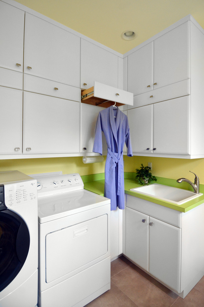 Modern inredning av en mellanstor l-formad tvättstuga enbart för tvätt, med en nedsänkt diskho, luckor med upphöjd panel, vita skåp, klinkergolv i porslin, en tvättmaskin och torktumlare bredvid varandra, brunt golv och gula väggar