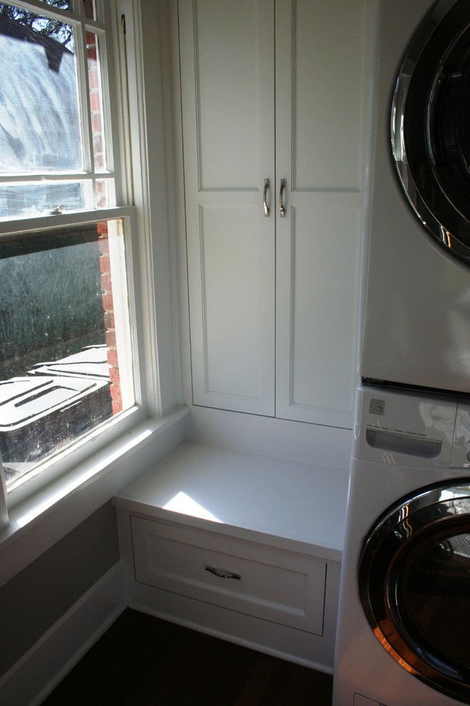 Kleine Klassische Waschküche in L-Form mit Schrankfronten im Shaker-Stil, weißen Schränken, weißer Wandfarbe, dunklem Holzboden und Waschmaschine und Trockner gestapelt in Dallas