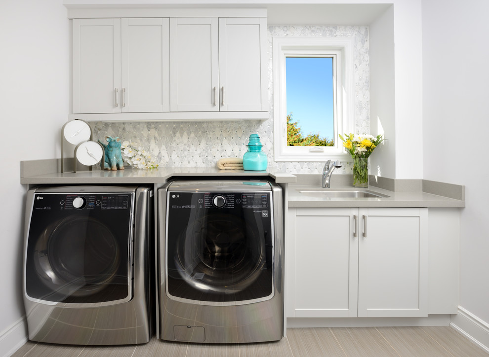 Exempel på en stor klassisk grå linjär grått tvättstuga enbart för tvätt, med en undermonterad diskho, luckor med infälld panel, grå skåp, bänkskiva i kvarts, grå väggar, klinkergolv i porslin, en tvättmaskin och torktumlare bredvid varandra och beiget golv