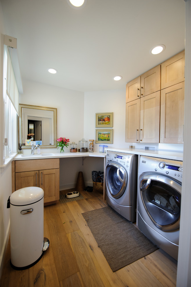 Inspiration for a mid-sized contemporary medium tone wood floor dedicated laundry room remodel in Indianapolis with an undermount sink, shaker cabinets, light wood cabinets, solid surface countertops, white walls and a side-by-side washer/dryer