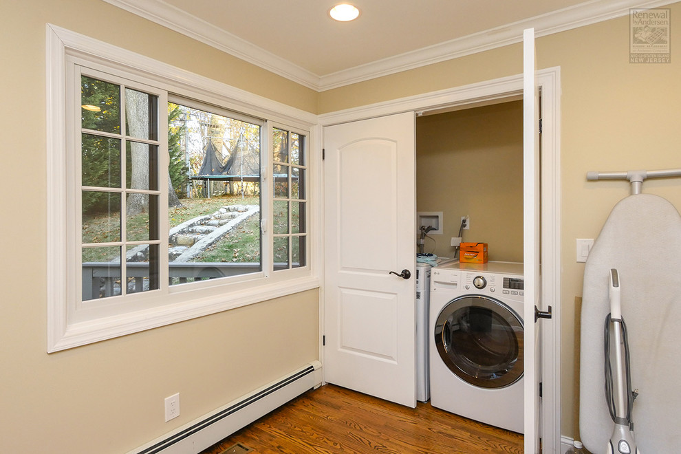 Modelo de cuarto de lavado de tamaño medio con paredes amarillas, suelo de madera en tonos medios, lavadora y secadora juntas y suelo marrón