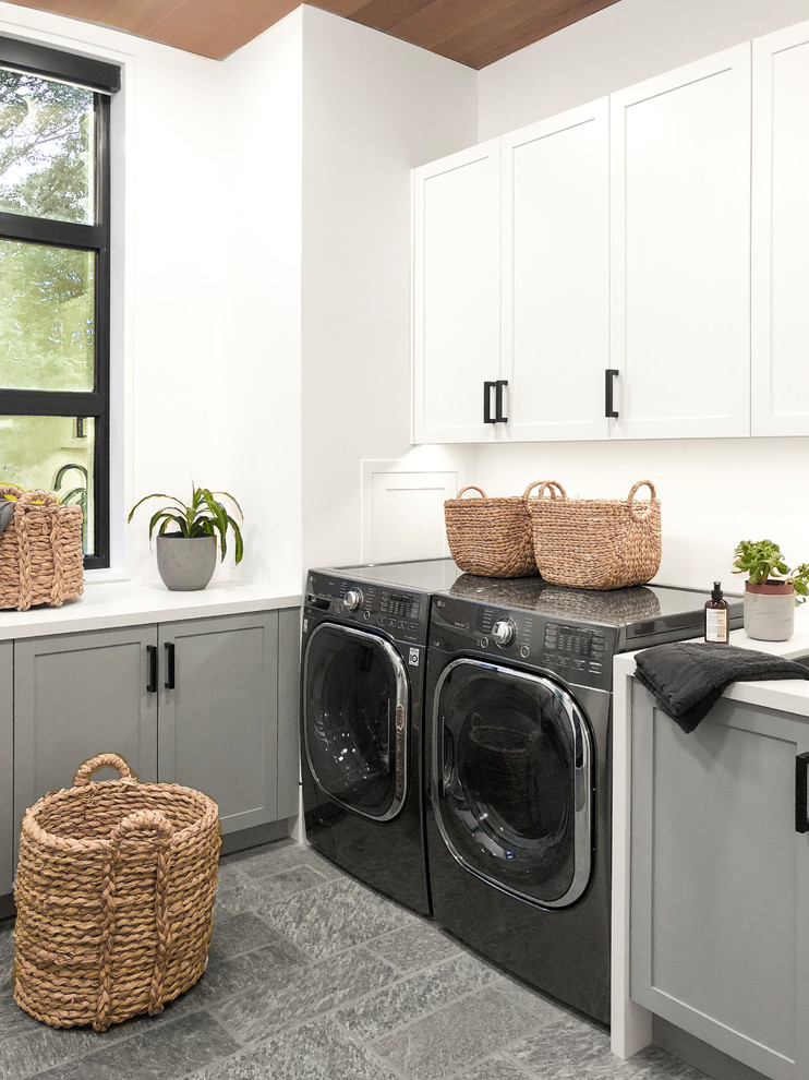 Inredning av en lantlig vita vitt tvättstuga enbart för tvätt, med skåp i shakerstil, grå skåp, vita väggar, en tvättmaskin och torktumlare bredvid varandra och grått golv