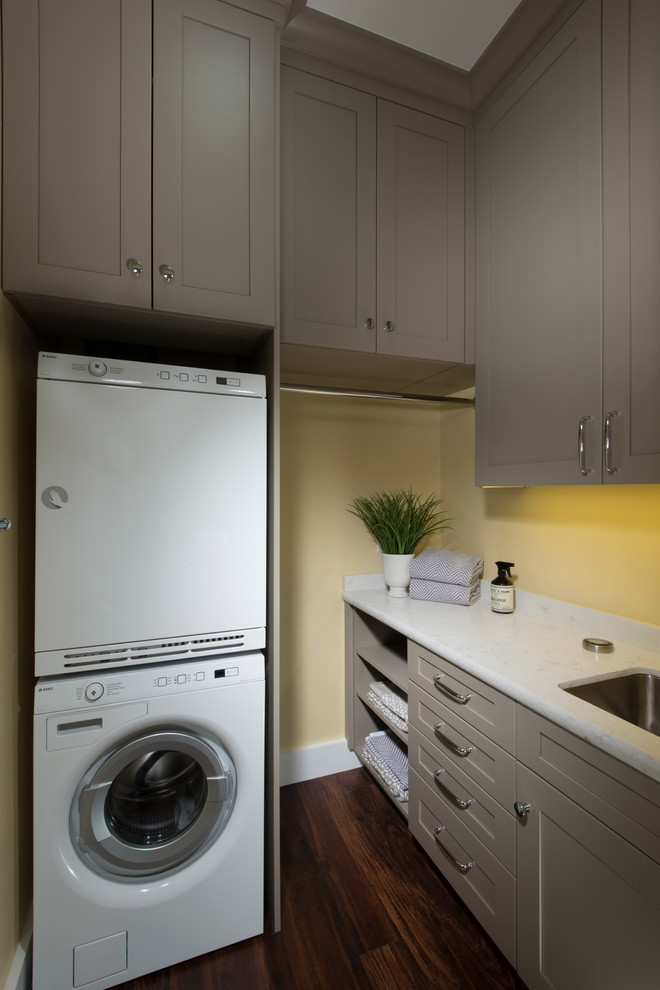 Foto på en liten amerikansk l-formad tvättstuga enbart för tvätt, med en undermonterad diskho, luckor med infälld panel, grå skåp, bänkskiva i kvarts, gula väggar, vinylgolv, en tvättpelare och brunt golv