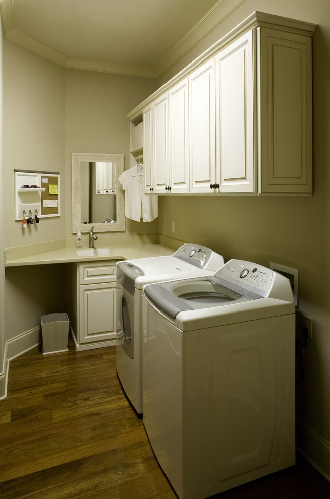 Idéer för mellanstora vintage l-formade beige tvättstugor enbart för tvätt, med en nedsänkt diskho, luckor med upphöjd panel, vita skåp, beige väggar, mellanmörkt trägolv, en tvättmaskin och torktumlare bredvid varandra, bänkskiva i kvarts och brunt golv