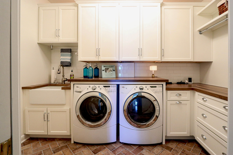 Example of a small transitional l-shaped porcelain tile and brown floor dedicated laundry room design in Cleveland with a farmhouse sink, flat-panel cabinets, white cabinets, wood countertops, white backsplash, ceramic backsplash, white walls, a side-by-side washer/dryer and brown countertops