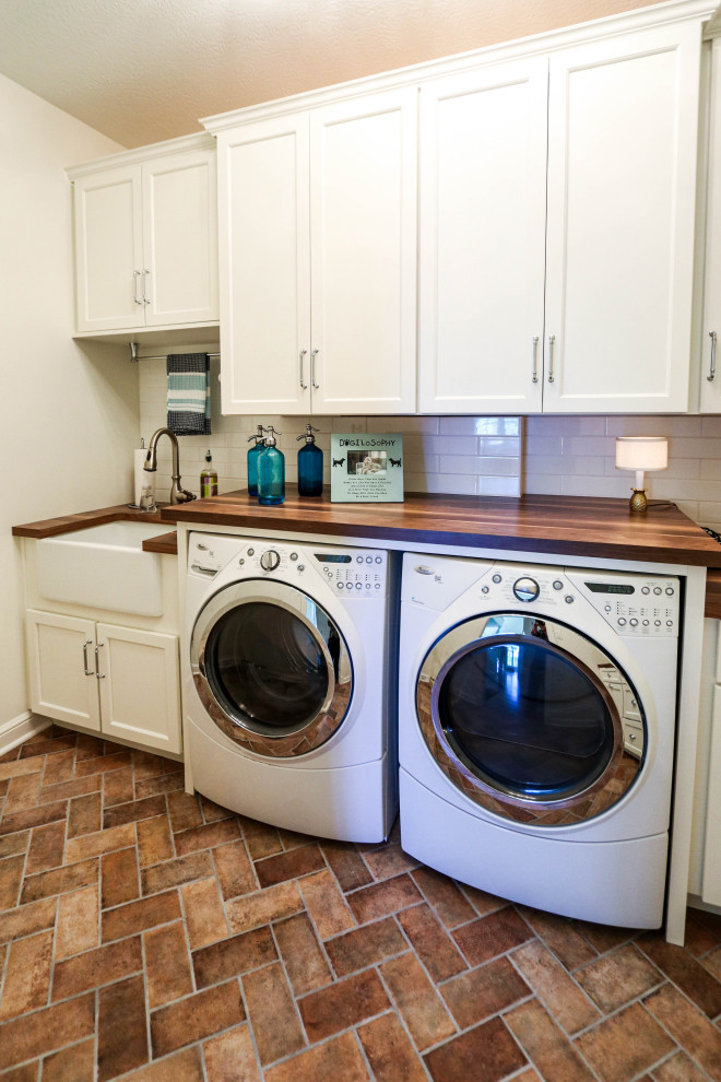 Dedicated laundry room - small transitional l-shaped porcelain tile and brown floor dedicated laundry room idea in Cleveland with a farmhouse sink, flat-panel cabinets, white cabinets, wood countertops, white backsplash, ceramic backsplash, white walls, a side-by-side washer/dryer and brown countertops