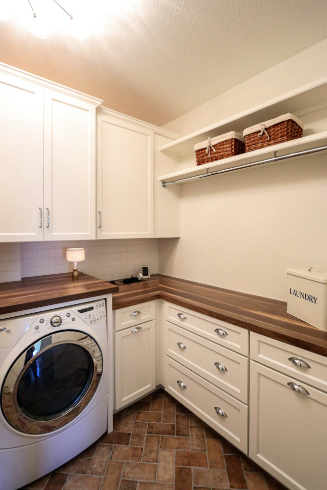 Exempel på en liten klassisk bruna l-formad brunt tvättstuga enbart för tvätt, med en rustik diskho, släta luckor, vita skåp, träbänkskiva, vitt stänkskydd, stänkskydd i keramik, vita väggar, klinkergolv i porslin, en tvättmaskin och torktumlare bredvid varandra och brunt golv
