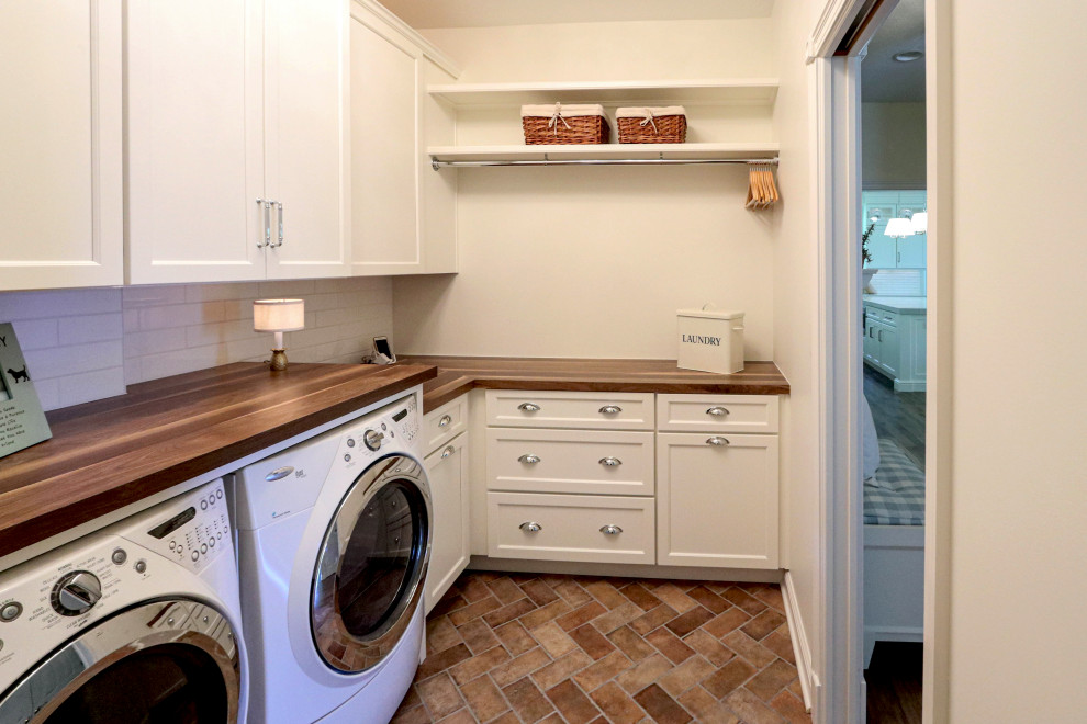 Idéer för små vintage l-formade brunt tvättstugor enbart för tvätt, med en rustik diskho, släta luckor, vita skåp, träbänkskiva, vitt stänkskydd, stänkskydd i keramik, vita väggar, klinkergolv i porslin, en tvättmaskin och torktumlare bredvid varandra och brunt golv