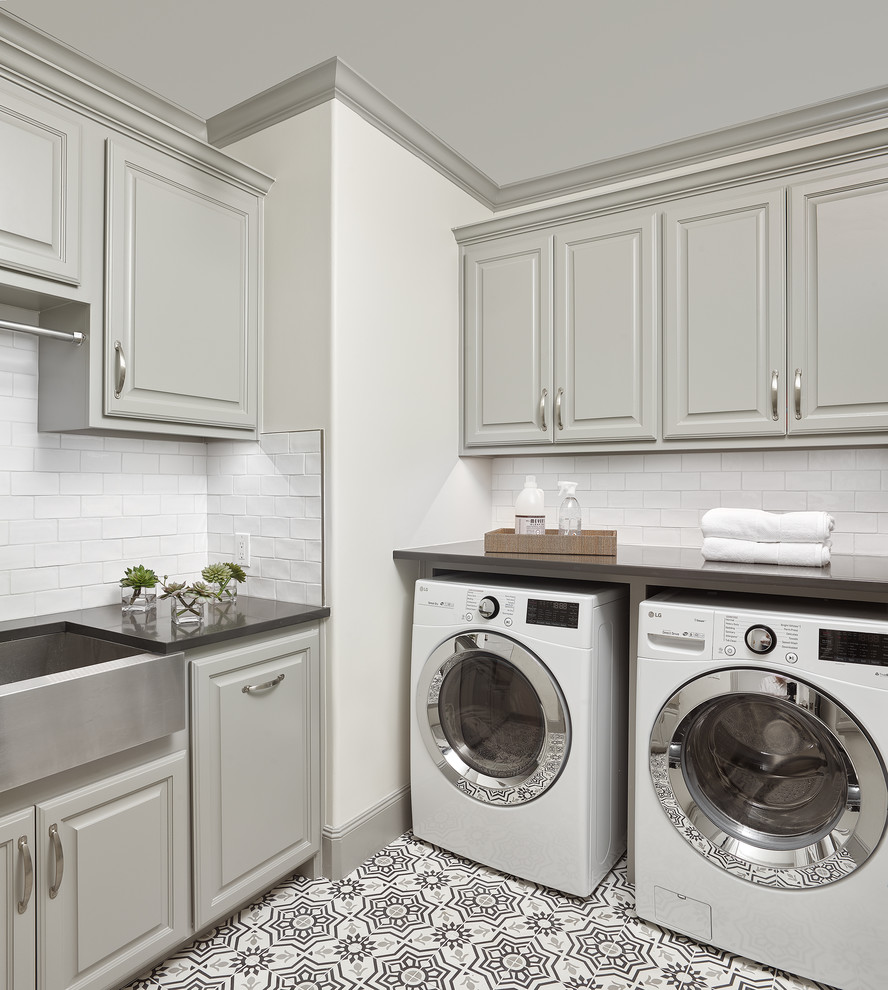 Klassisk inredning av en bruna l-formad brunt tvättstuga enbart för tvätt, med luckor med upphöjd panel, grå skåp, vita väggar, en tvättmaskin och torktumlare bredvid varandra, flerfärgat golv och vitt stänkskydd