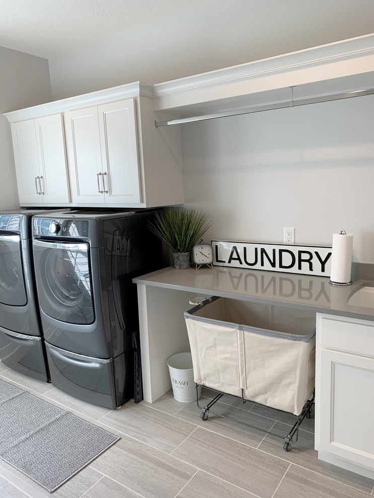 Exempel på en mellanstor klassisk grå linjär grått tvättstuga enbart för tvätt, med en undermonterad diskho, släta luckor, vita skåp, bänkskiva i kvarts, klinkergolv i keramik, en tvättmaskin och torktumlare bredvid varandra och grått golv