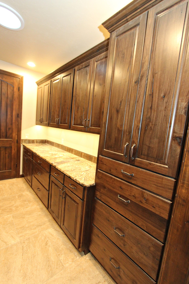 Photo of a large classic galley separated utility room in Boise with a submerged sink, raised-panel cabinets, dark wood cabinets, granite worktops, beige walls, travertine flooring and a stacked washer and dryer.