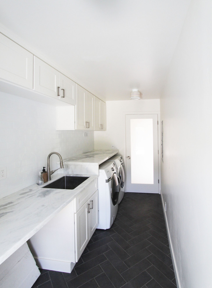 Inredning av en liten grå parallell grått tvättstuga enbart för tvätt, med en undermonterad diskho, skåp i shakerstil, vita skåp, bänkskiva i kvartsit, vita väggar, klinkergolv i porslin, en tvättmaskin och torktumlare bredvid varandra och svart golv