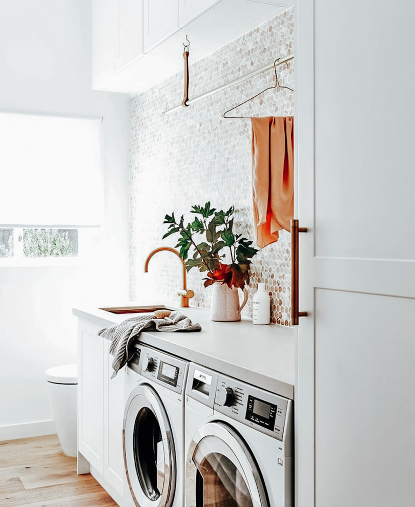 Idéer för en mellanstor grå linjär tvättstuga enbart för tvätt, med en undermonterad diskho, vita skåp, bänkskiva i kvartsit, grått stänkskydd, stänkskydd i mosaik, vita väggar, mellanmörkt trägolv, en tvättmaskin och torktumlare bredvid varandra och brunt golv