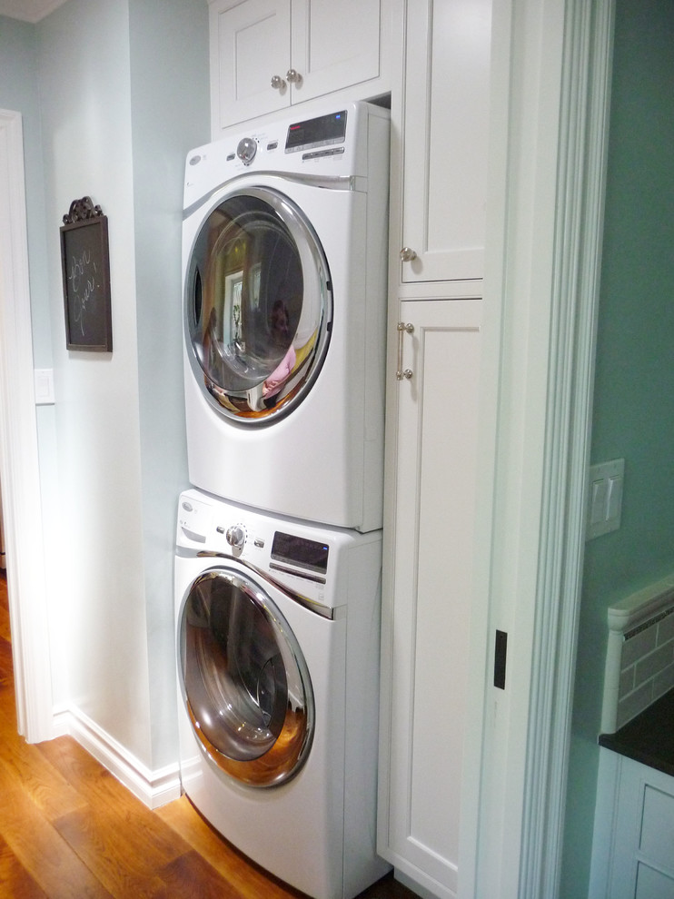 Modelo de lavadero clásico de tamaño medio con armarios con paneles empotrados, puertas de armario blancas, paredes azules, suelo de madera en tonos medios y lavadora y secadora apiladas