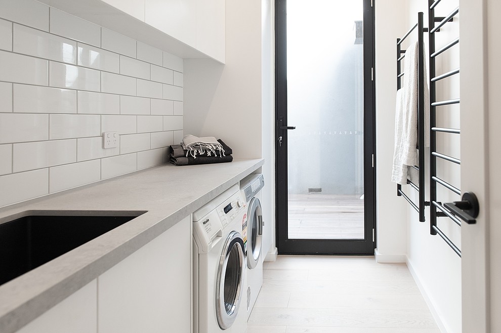 Idéer för mellanstora funkis linjära grått tvättstugor enbart för tvätt, med en undermonterad diskho, vita skåp, bänkskiva i kvarts, vita väggar, ljust trägolv, en tvättmaskin och torktumlare bredvid varandra, släta luckor och beiget golv
