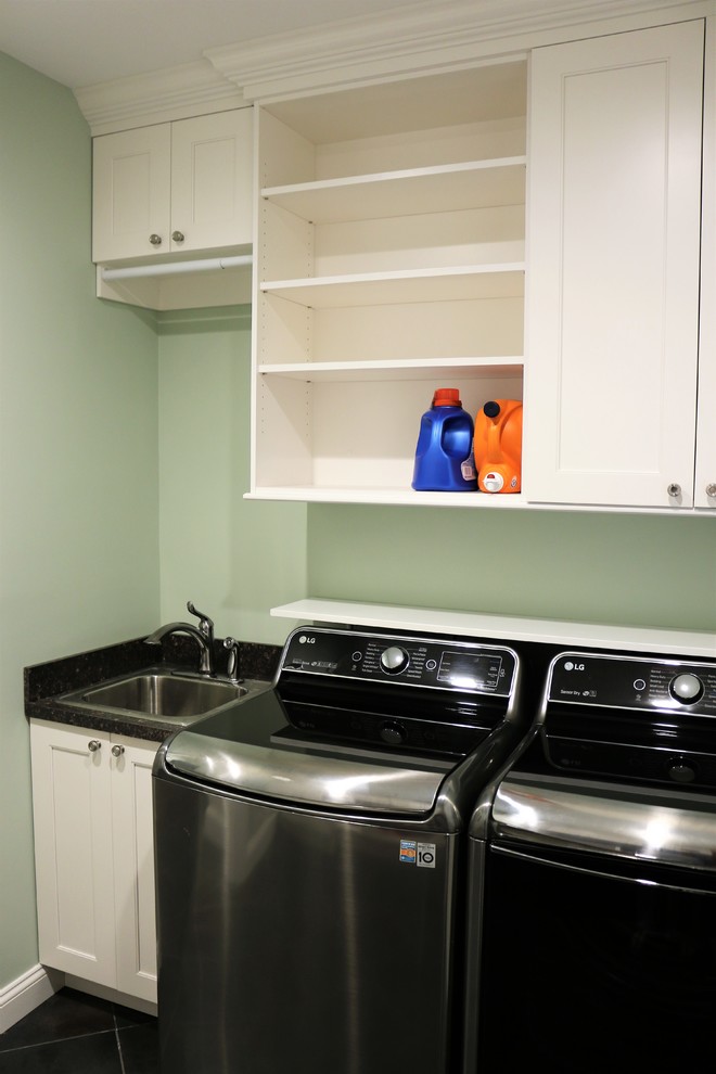 Lantlig inredning av en mellanstor parallell tvättstuga enbart för tvätt, med en nedsänkt diskho, öppna hyllor, vita skåp, laminatbänkskiva, gröna väggar, klinkergolv i porslin, en tvättmaskin och torktumlare bredvid varandra och svart golv
