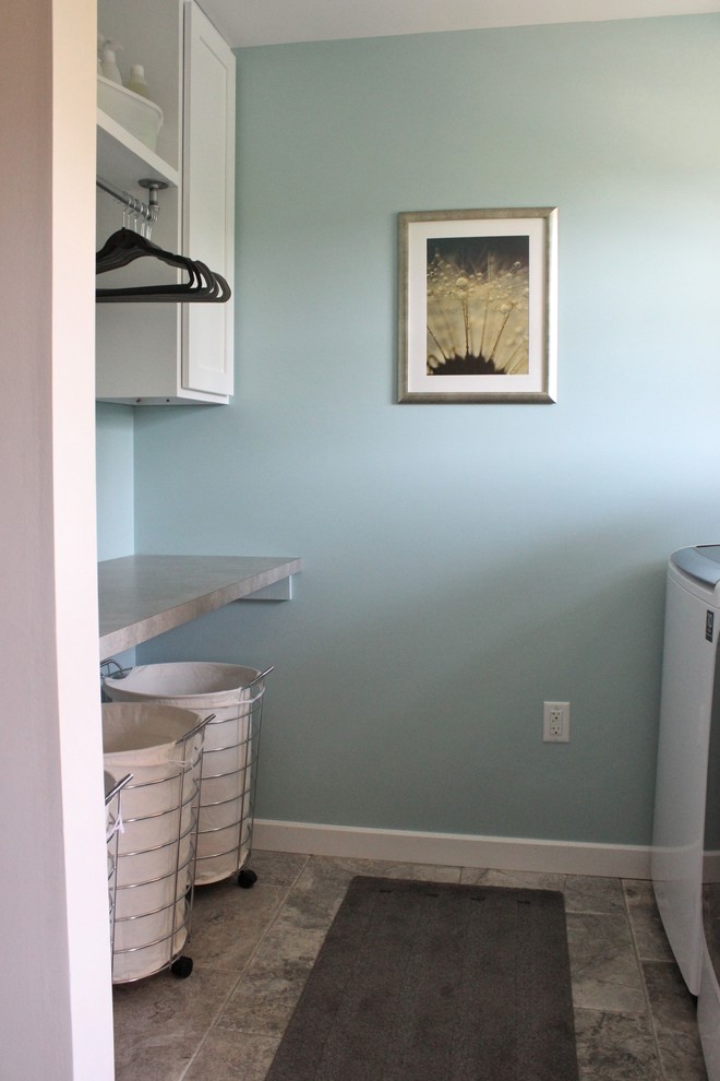 Modelo de cuarto de lavado de galera industrial con armarios estilo shaker, puertas de armario blancas, encimera de laminado, paredes azules, suelo de travertino y lavadora y secadora juntas