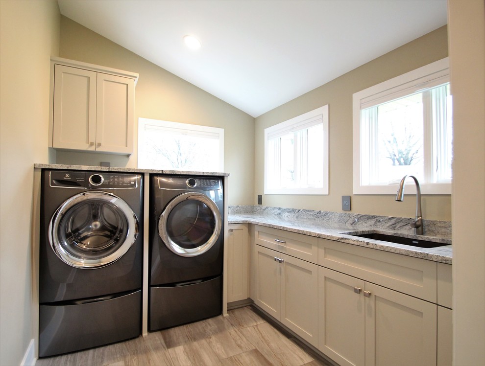 Exempel på en stor klassisk flerfärgade l-formad flerfärgat tvättstuga enbart för tvätt, med en undermonterad diskho, skåp i shakerstil, vita skåp, granitbänkskiva, klinkergolv i porslin, en tvättmaskin och torktumlare bredvid varandra, grått golv och beige väggar