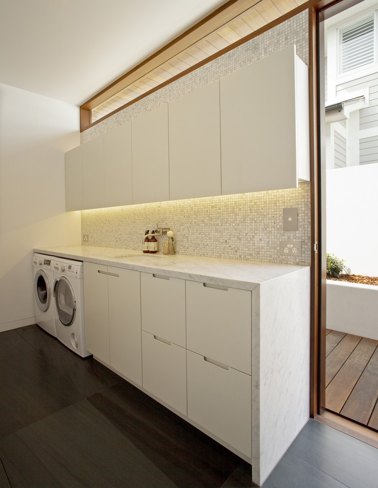 Exempel på en stor modern vita vitt tvättstuga enbart för tvätt, med en undermonterad diskho, vita skåp, marmorbänkskiva, grå väggar, skiffergolv och en tvättmaskin och torktumlare bredvid varandra
