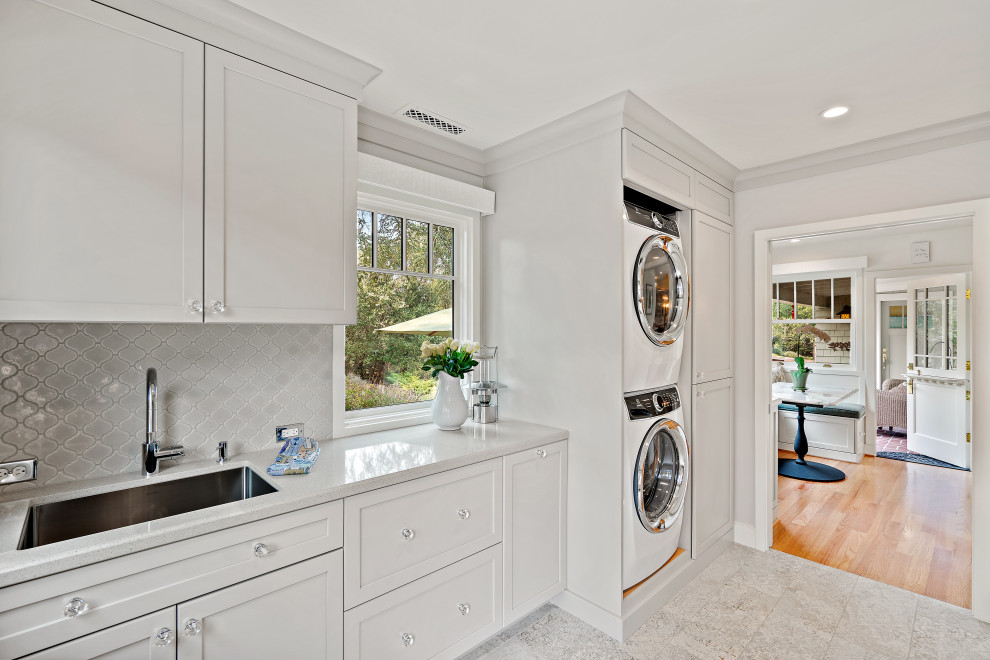 Mid-sized elegant single-wall ceramic tile and gray floor utility room photo in San Francisco with an undermount sink, shaker cabinets, white cabinets, quartz countertops, gray backsplash, ceramic backsplash, white walls, a stacked washer/dryer and white countertops