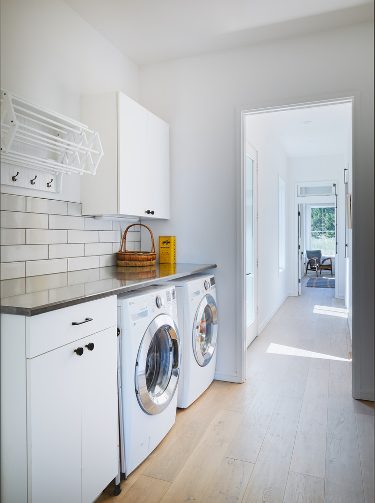 Immagine di una sala lavanderia nordica con ante lisce, ante bianche, top in acciaio inossidabile, pareti bianche, parquet chiaro, lavatrice e asciugatrice affiancate, pavimento beige e top grigio