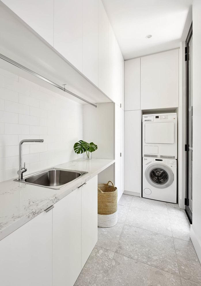 Diseño de lavadero en L moderno con fregadero encastrado, armarios con paneles lisos, puertas de armario blancas, lavadora y secadora apiladas, suelo gris y encimeras blancas