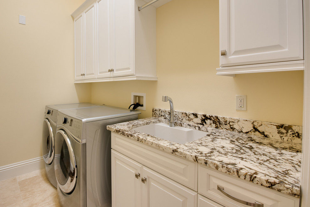 Inredning av en klassisk linjär tvättstuga enbart för tvätt, med en undermonterad diskho, vita skåp, granitbänkskiva, travertin golv och en tvättmaskin och torktumlare bredvid varandra