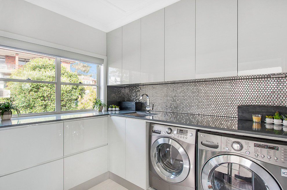 Moderne Waschküche mit weißen Schränken, Granit-Arbeitsplatte, Waschmaschine und Trockner nebeneinander, Unterbauwaschbecken, flächenbündigen Schrankfronten und grauer Arbeitsplatte in Newcastle - Maitland