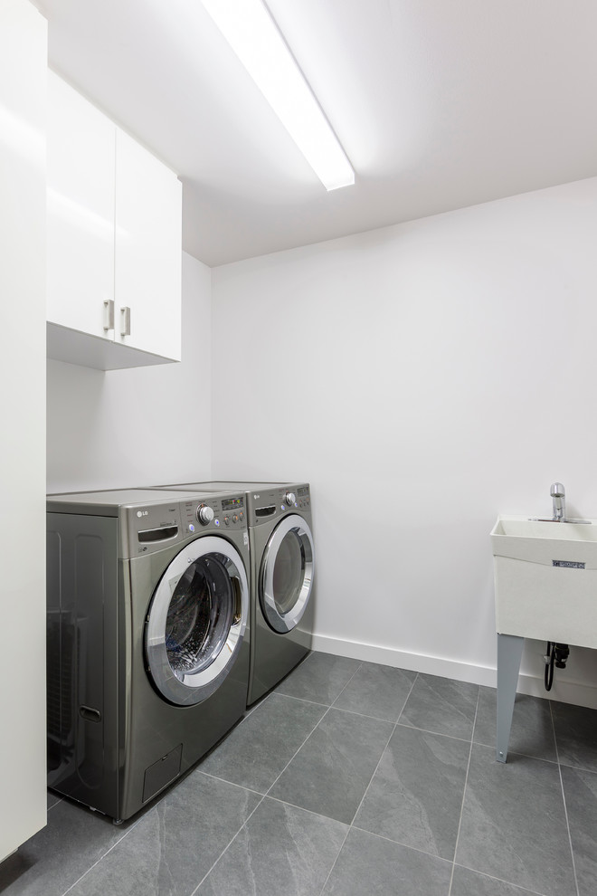 Foto på en mellanstor funkis linjär tvättstuga enbart för tvätt, med en allbänk, släta luckor, vita skåp, vita väggar, klinkergolv i keramik, en tvättmaskin och torktumlare bredvid varandra och grått golv