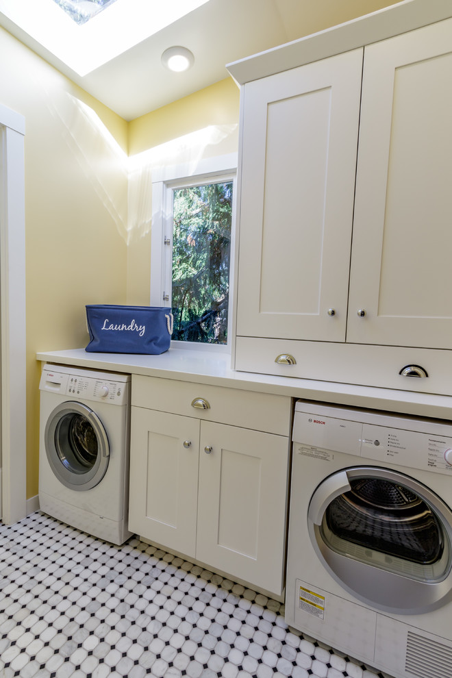 Inredning av en amerikansk mellanstor parallell tvättstuga enbart för tvätt, med skåp i shakerstil, vita skåp, bänkskiva i koppar, gula väggar, marmorgolv och en tvättmaskin och torktumlare bredvid varandra