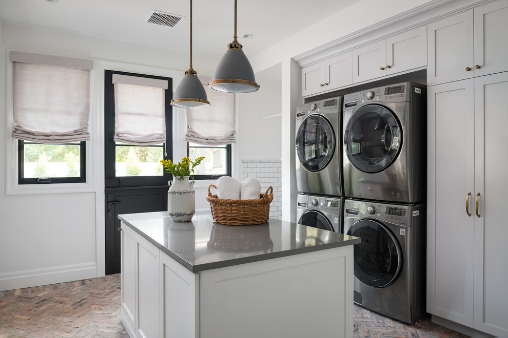 Foto de lavadero clásico renovado con armarios estilo shaker, puertas de armario grises, paredes blancas, suelo de ladrillo, lavadora y secadora apiladas, suelo rojo y encimeras grises
