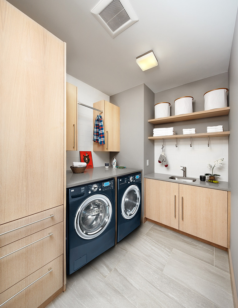 Modern inredning av en grå l-formad grått tvättstuga, med en undermonterad diskho, släta luckor, skåp i ljust trä, grå väggar, en tvättmaskin och torktumlare bredvid varandra och grått golv
