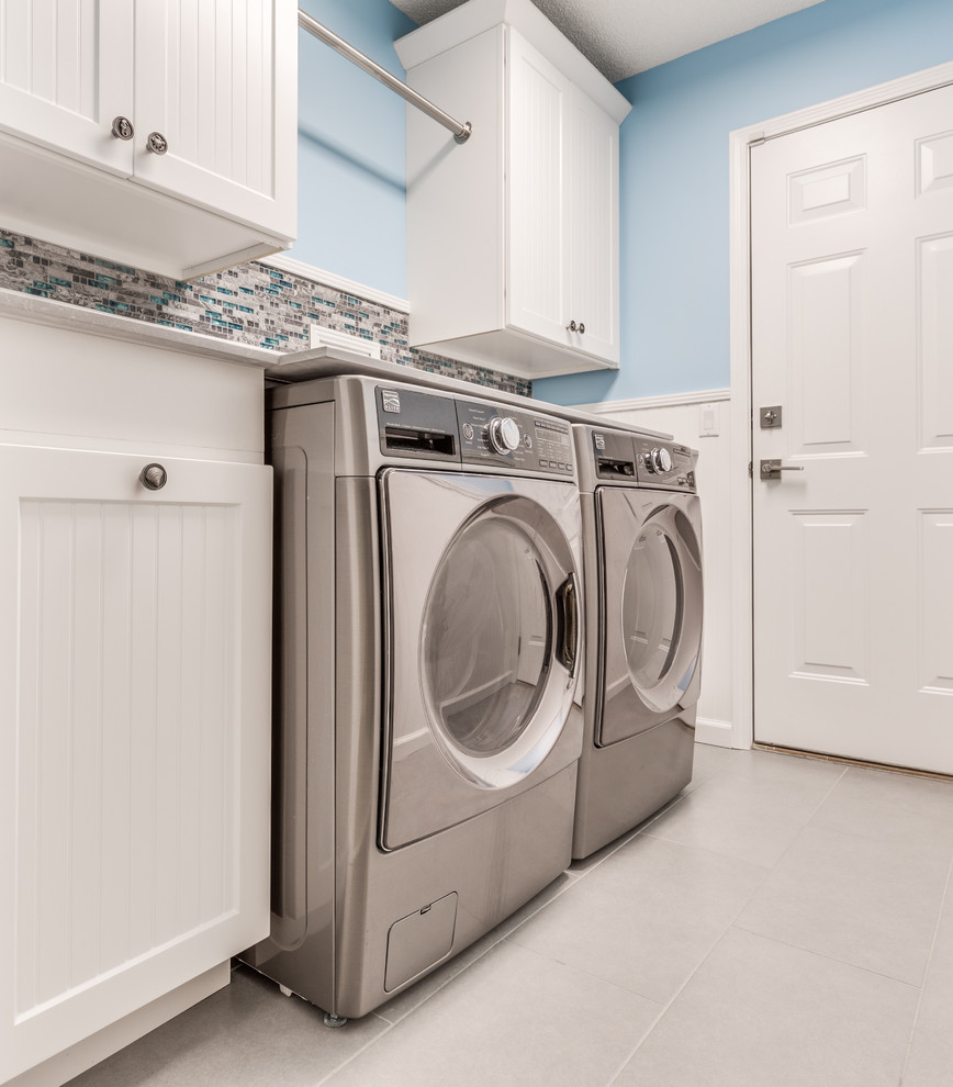 Klassisk inredning av en mellanstor linjär tvättstuga enbart för tvätt, med luckor med infälld panel, vita skåp, marmorbänkskiva, blå väggar, klinkergolv i porslin och en tvättmaskin och torktumlare bredvid varandra