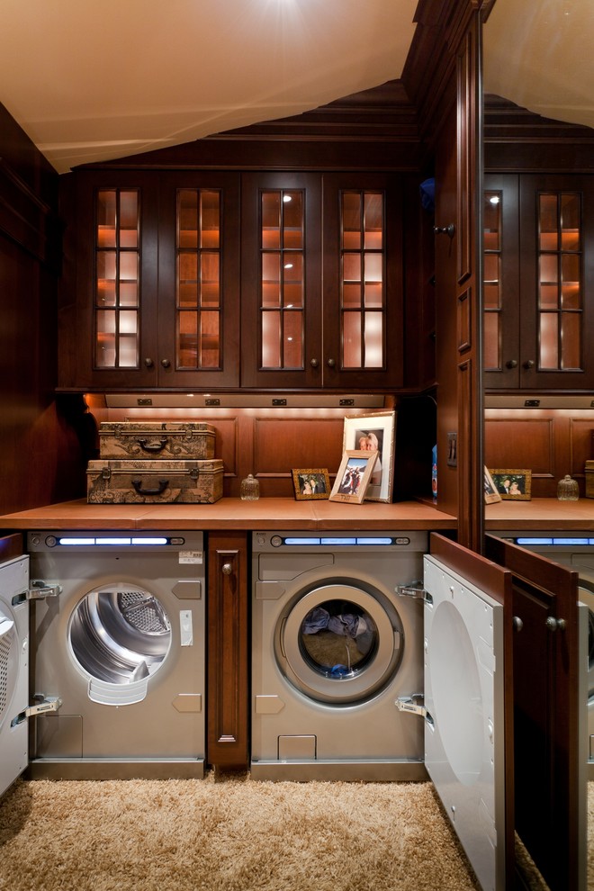 This is an example of an expansive traditional utility room in Boston with glass-front cabinets, dark wood cabinets, carpet, a side by side washer and dryer, beige floors and brown worktops.