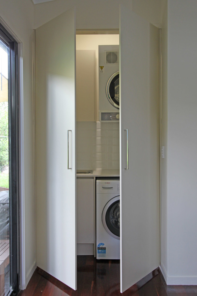 This is an example of a small contemporary single-wall separated utility room in Brisbane with a built-in sink, flat-panel cabinets, white cabinets, engineered stone countertops, white walls, dark hardwood flooring, a stacked washer and dryer, brown floors and beige worktops.