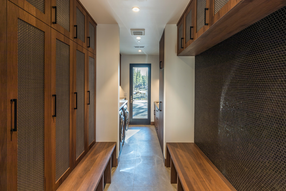 Cette image montre une buanderie parallèle design en bois brun multi-usage avec un évier encastré, un placard à porte shaker, un mur blanc, des machines côte à côte, un sol gris et un plan de travail gris.