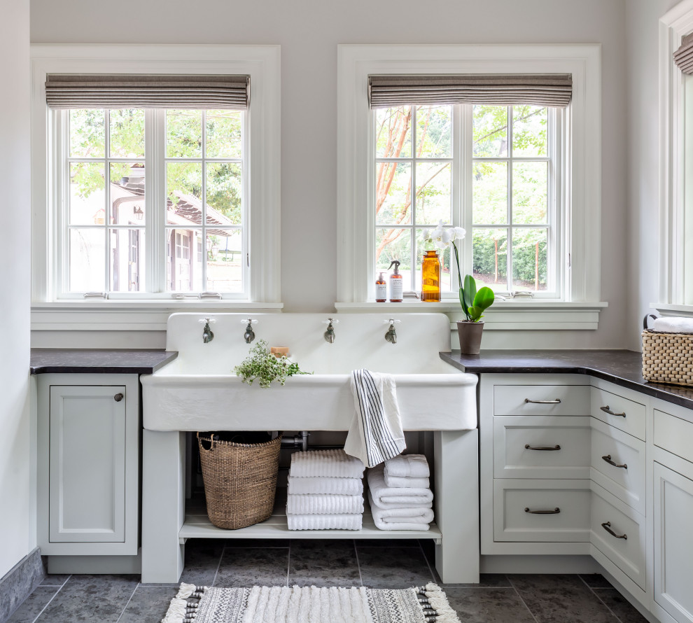 Klassische Waschküche in L-Form mit Landhausspüle, Schrankfronten mit vertiefter Füllung, weißen Schränken, grauer Wandfarbe, grauem Boden und schwarzer Arbeitsplatte in Atlanta