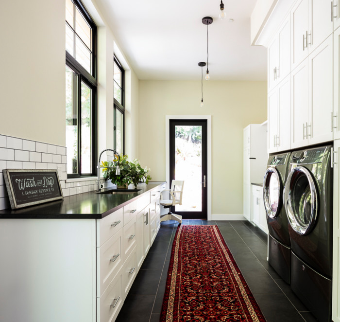 Foto på en stor lantlig svarta parallell tvättstuga enbart för tvätt, med en undermonterad diskho, skåp i shakerstil, vita skåp, granitbänkskiva, gula väggar, klinkergolv i porslin, en tvättmaskin och torktumlare bredvid varandra och svart golv