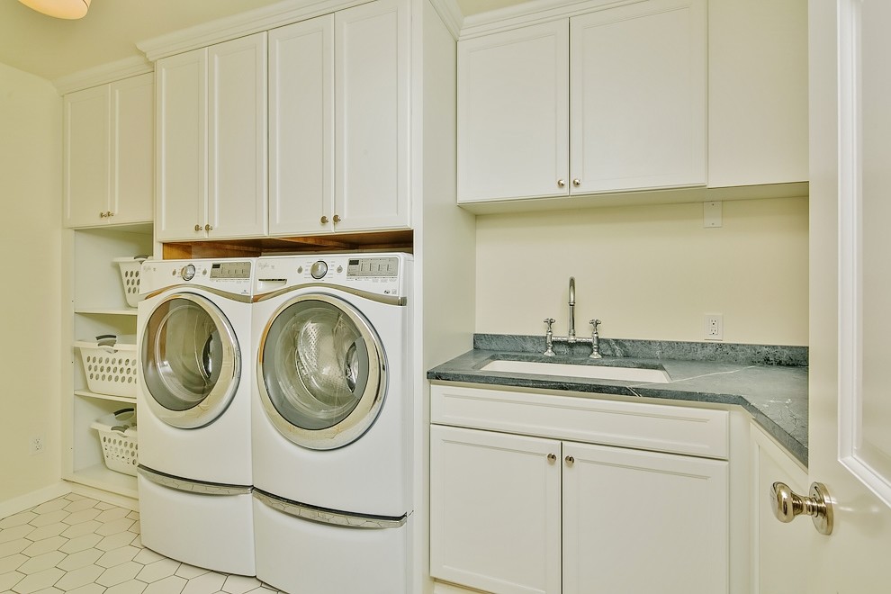 Klassisk inredning av en stor l-formad tvättstuga enbart för tvätt, med en undermonterad diskho, skåp i shakerstil, vita skåp, vita väggar, laminatgolv och en tvättmaskin och torktumlare bredvid varandra