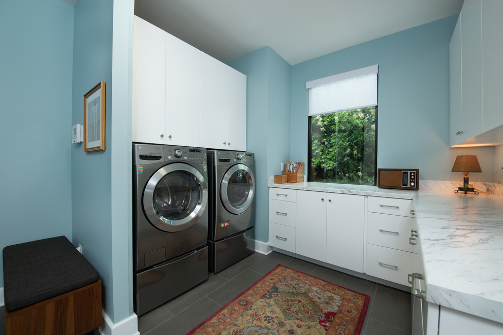 Kleine Mid-Century Waschküche in U-Form mit flächenbündigen Schrankfronten, weißen Schränken, Laminat-Arbeitsplatte, blauer Wandfarbe, Porzellan-Bodenfliesen, Waschmaschine und Trockner nebeneinander, grauem Boden und weißer Arbeitsplatte in Atlanta