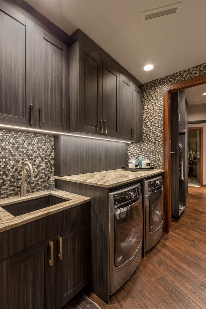Example of a small trendy single-wall dark wood floor and brown floor dedicated laundry room design in Omaha with an undermount sink, recessed-panel cabinets, gray cabinets, granite countertops, a side-by-side washer/dryer and multicolored countertops