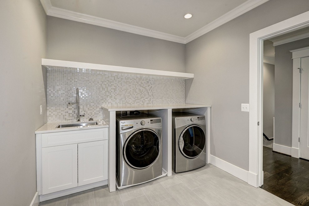 Inredning av en modern stor grå linjär grått tvättstuga enbart för tvätt, med en undermonterad diskho, skåp i shakerstil, vita skåp, bänkskiva i kvarts, grå väggar, klinkergolv i porslin, en tvättmaskin och torktumlare bredvid varandra och grått golv