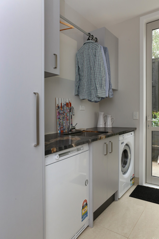Exempel på en liten modern linjär tvättstuga enbart för tvätt, med en undermonterad diskho, släta luckor, grå skåp, granitbänkskiva, grå väggar, klinkergolv i porslin, en tvättmaskin och torktumlare bredvid varandra och vitt golv