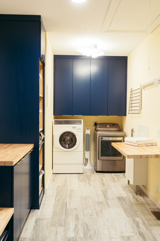 Esempio di una grande lavanderia multiuso minimalista con lavello a vasca singola, ante in stile shaker, ante blu, top in legno, pareti gialle, pavimento in gres porcellanato, lavatrice e asciugatrice affiancate, pavimento grigio e top marrone
