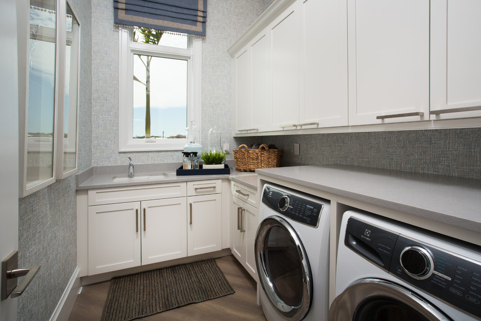 Inredning av en mellanstor grå grått tvättstuga enbart för tvätt, med en nedsänkt diskho, vita skåp, blå väggar, mellanmörkt trägolv, en tvättmaskin och torktumlare bredvid varandra och beiget golv