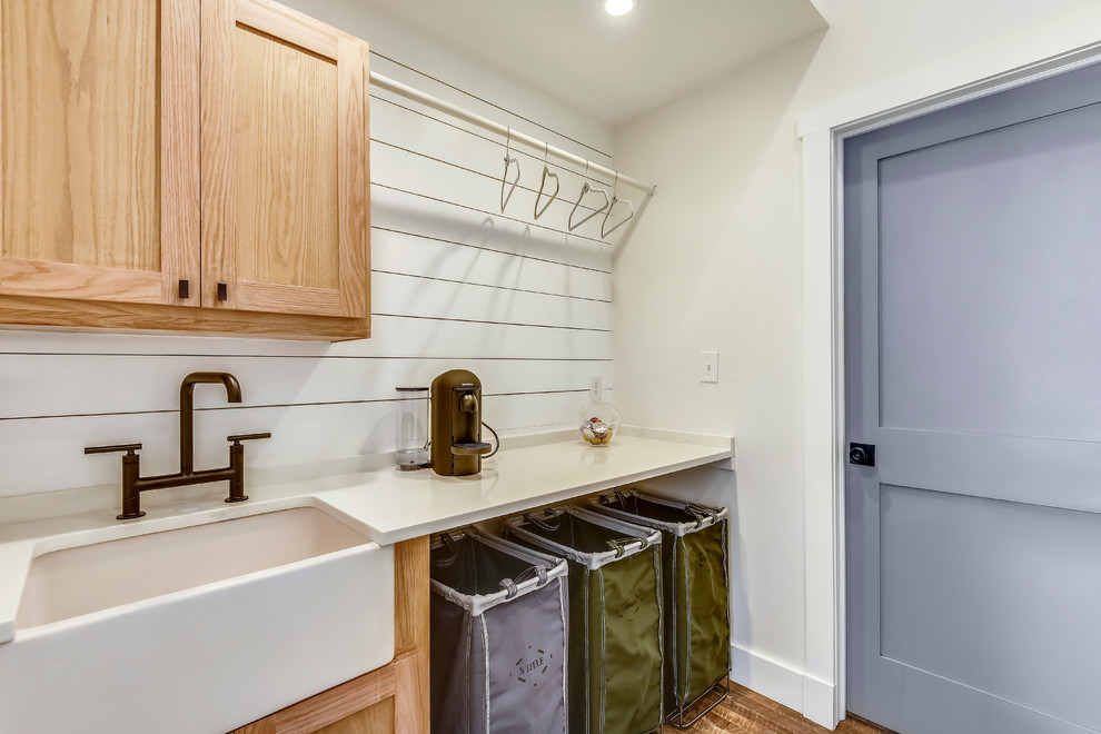 Photo of a medium sized farmhouse single-wall separated utility room in DC Metro with a belfast sink, shaker cabinets, light wood cabinets, quartz worktops, white walls, medium hardwood flooring, a stacked washer and dryer, brown floors and white worktops.